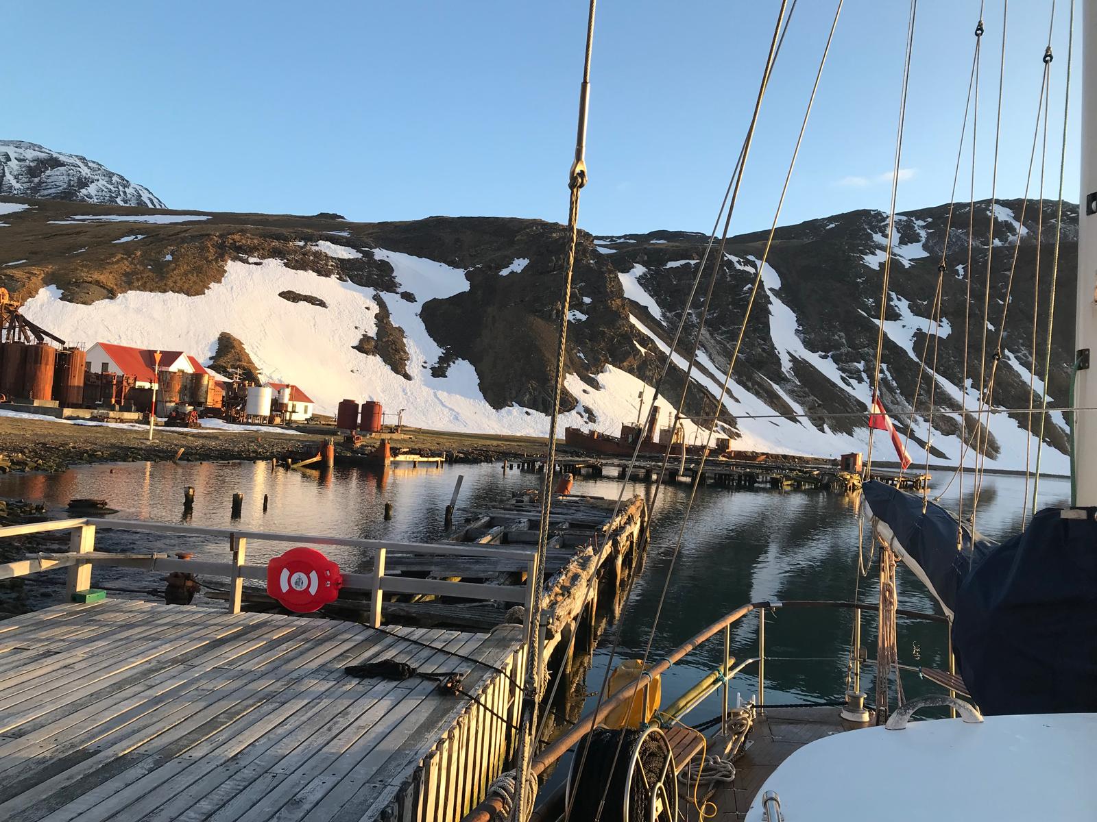 Alongside at the old jetty in evening light. Oil tanks and red-roofed buildings ashore, a hulk beyond the pontoon.