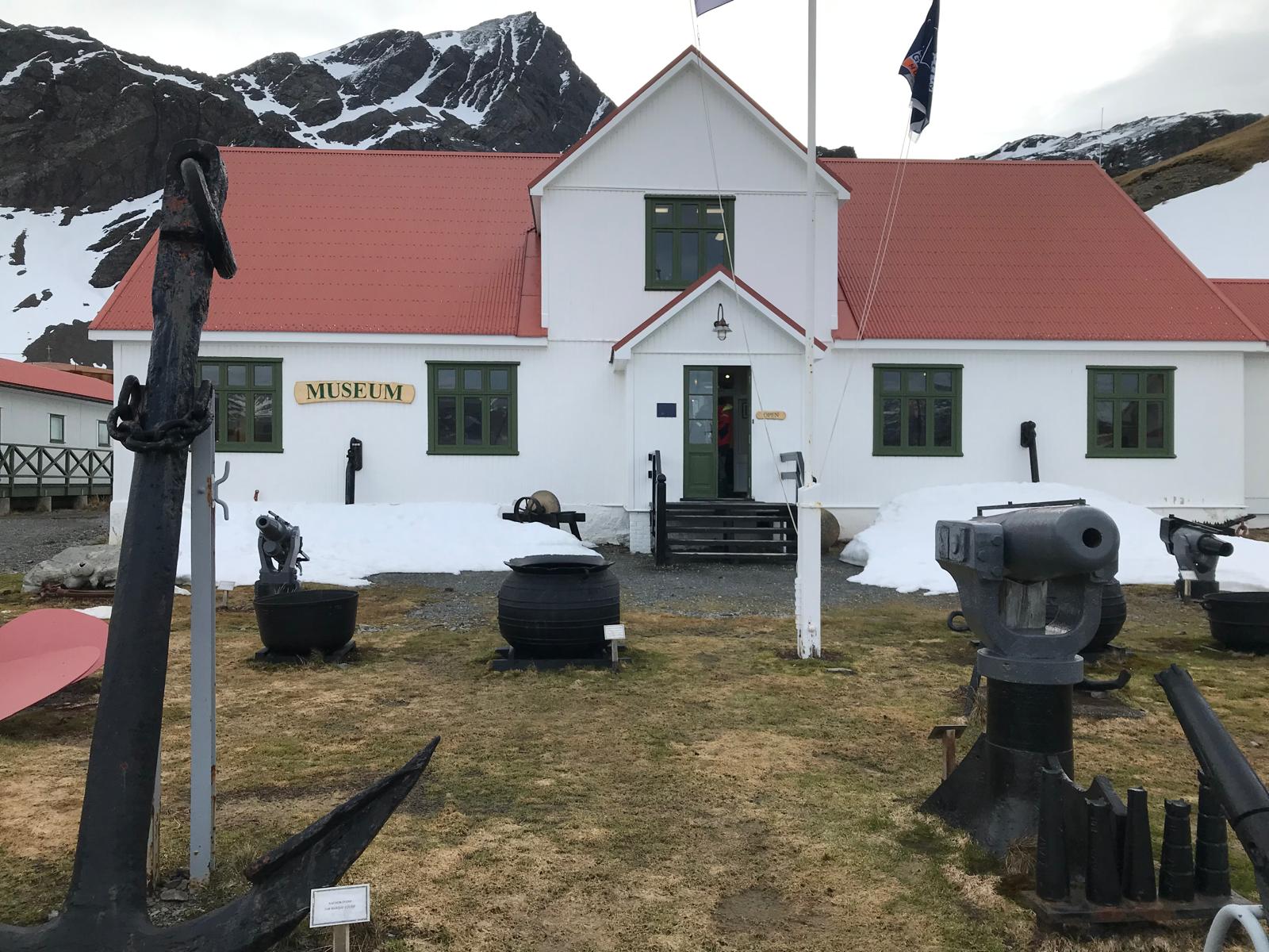 The South Georgia Museum — white weatherboard, red roof, harpoon guns and try-pots on the grass outside, an anchor in the foreground.