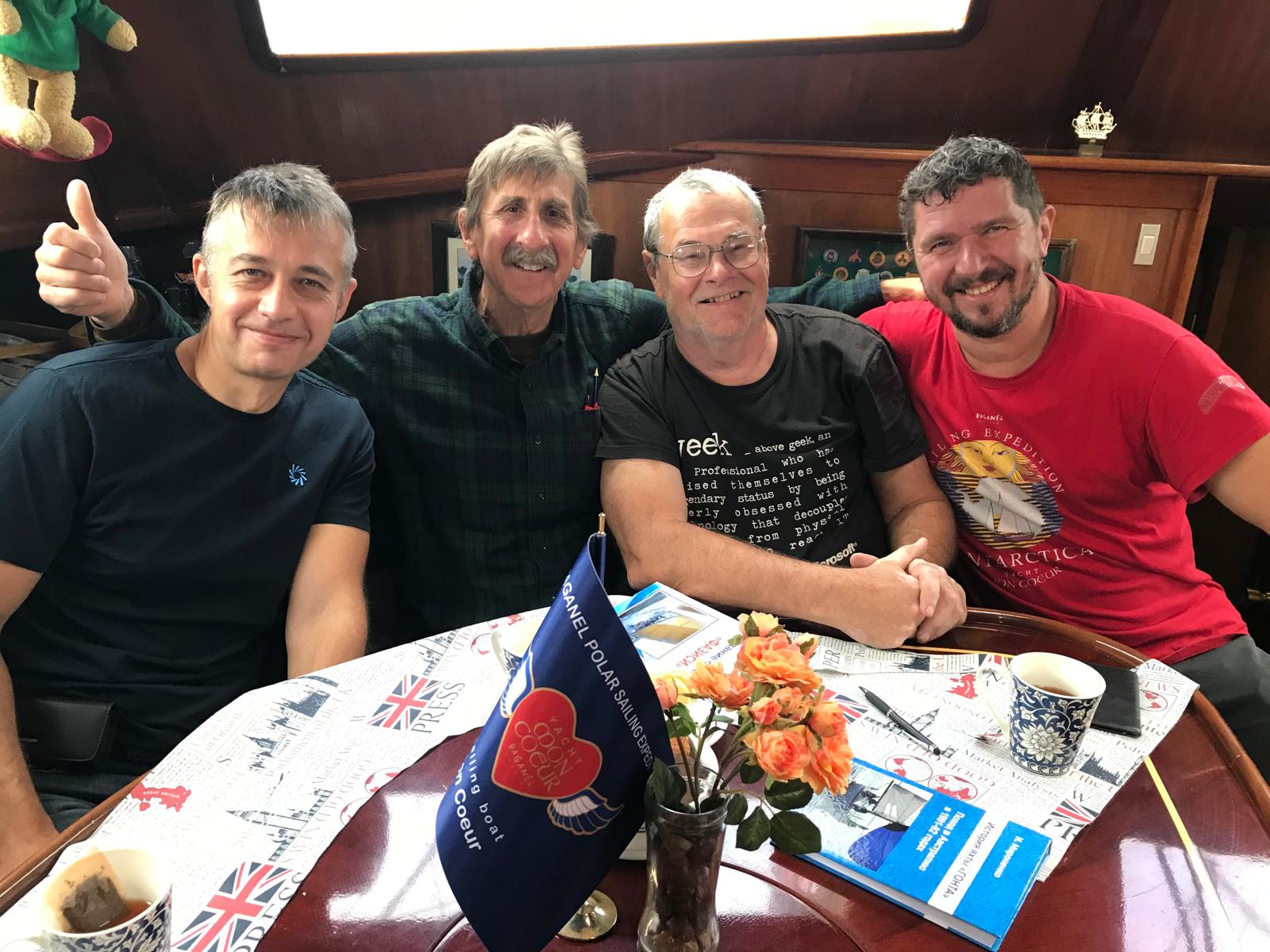 Four of the crew round the saloon table. The table flag reads Paganel Polar Sailing Expedition, sailing boat Mon Coeur; one shirt reads Paganel Sailing Expedition Antarctica.